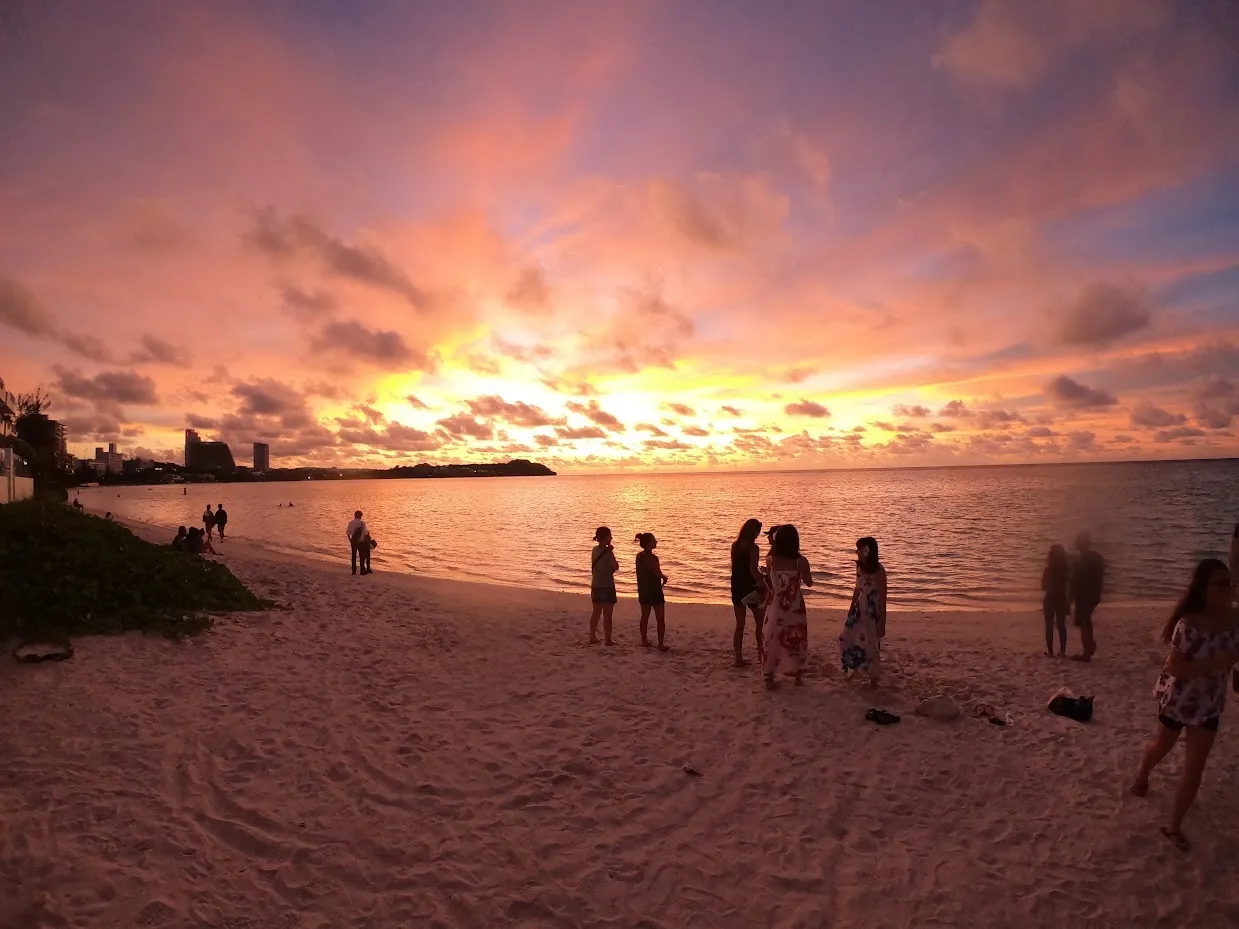 Sunset over Tumon Bay, Guam. The sky is orange and violet, and people stand along the beach in silhouette.